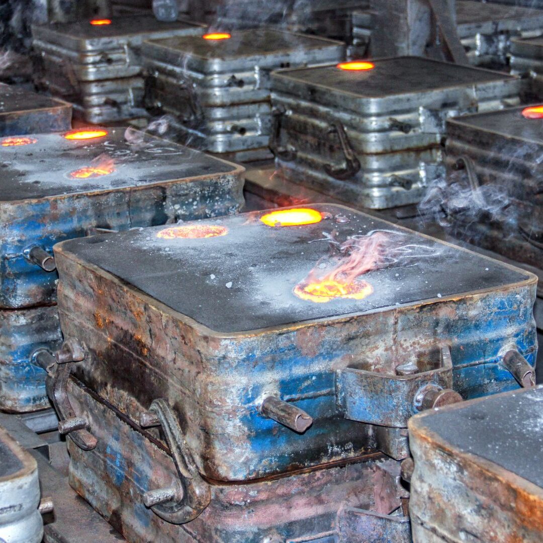A foundry scene showing multiple sand casting molds with molten metal being poured, glowing with heat and emitting smoke during the solidification process.